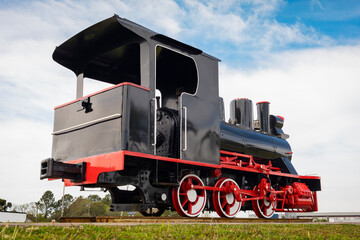 Historic black and red steam locomotive displayed outdoors on railway tracks beneath a dramatic blue and white sky. © MARCELO