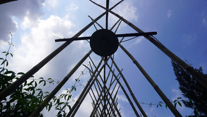 Low angle view of bamboo poles converging into tepee style frame with round wooden disc at center, silhouetted against blue sky with white clouds and climbing vines. © anungcamui