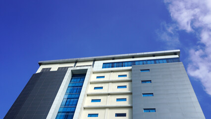 Modern multi story building with blue glass windows, gray panels and cream facade, rising against deep blue sky with white cloud drifting at the right edge. © anungcamui