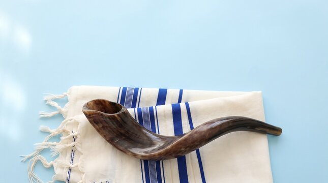 Brown shofar ram's horn resting on a white and blue striped tallit prayer shawl against a light blue background for Rosh Hashanah and Yom Kippur