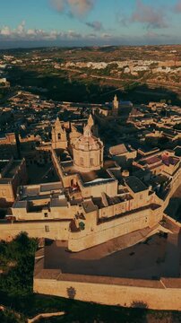 Vertical 9:16 drone view over the bastions of Mdina, Malta. Fortress walls, flat limestone rooftops and a church dome with long shadows in low golden hour light, countryside to the horizon.