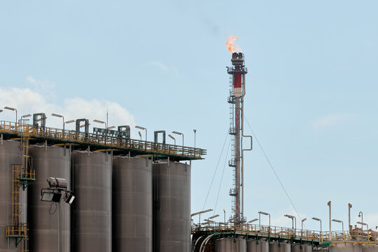 Industrial gas flare stack burning off excess gas above petrochemical refinery facility with rows of vertical stainless steel storage silos, catwalks, and floodlights under daylight sky with clouds