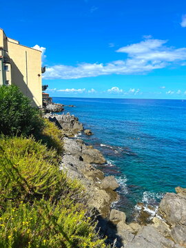 Scenic top-angle view of a Sicilian seascape with dramatic rocky cliffs and crags along the coastline. Crystal-clear turquoise Mediterranean waters crashing against the rugged shore under bright sunli