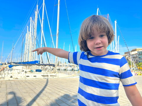 Portrait of a joyful smiling boy against a background of sailing boats. Yachting with children, family sea voyage. A child in a sailor t-shirt, ready to take the steering wheel