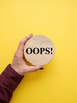 Hand Holding a Round Wooden Disc with Oops Text Over a Bright Yellow Background for Error Concept