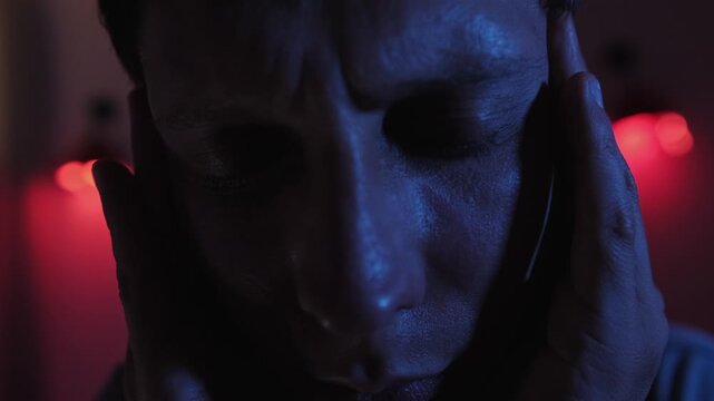 Man suffering from severe headache and migraine pain, pressing and massaging his temples with both hands in a dramatic extreme close up with blue face light and red background