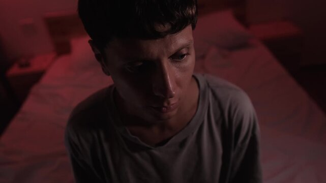 Man sitting alone and lost in rumination, looking downward while repeatedly replaying thoughts and memories in his mind, portraying mental fixation and unresolved personal conflict in a dark bedroom