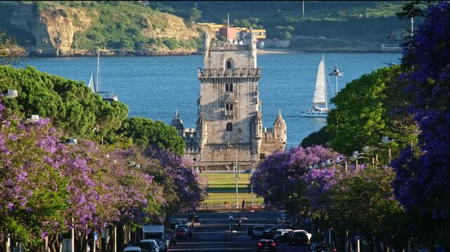 Blooming jacaranda trees frame Belem Tower in Belem, Lisbon, Portugal. Tourist boats sail across the water at sunset, creating a lively scene in the city.