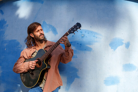 Musician playing acoustic guitar, strumming chords with passion, creating music on blue urban wall