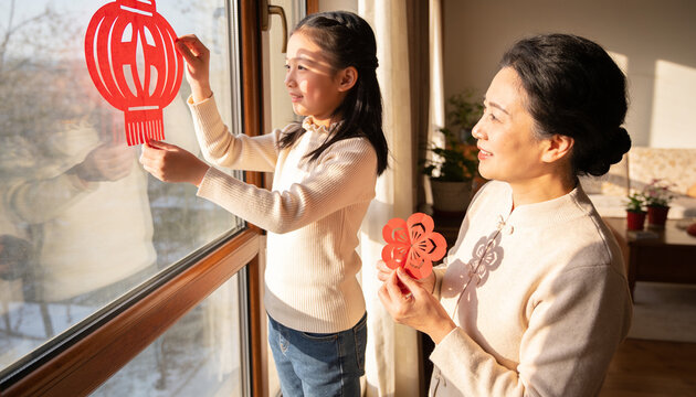 Asian mother and daughter decorating window with traditional red paper cut lanterns and flowers for Chinese New Year celebration at home in sunlight