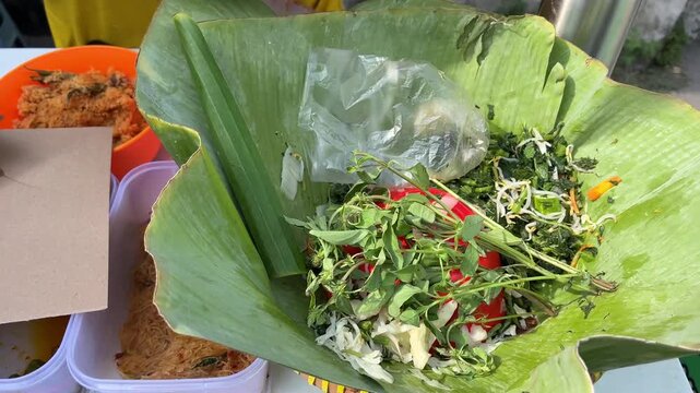 Vendor hand preparing traditional Indonesian pecel salad with fresh basil and boiled vegetables on banana leaf wrap, Asian street food.