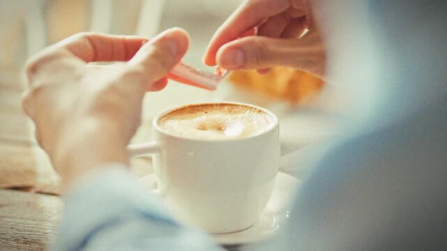 A woman's hands add sugar from a packet into a white ceramic cup of fresh cappuccino a croissant lies on a wooden table, with a sunlit caf&eacute; window in the background-a workday coffee break