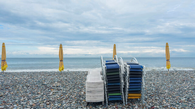 Pebble beach with stacked blue sun loungers and closed yellow striped umbrellas beside calm sea, cloudy sky, empty seaside resort in off season, travel destination, summer holiday, coastal relaxation