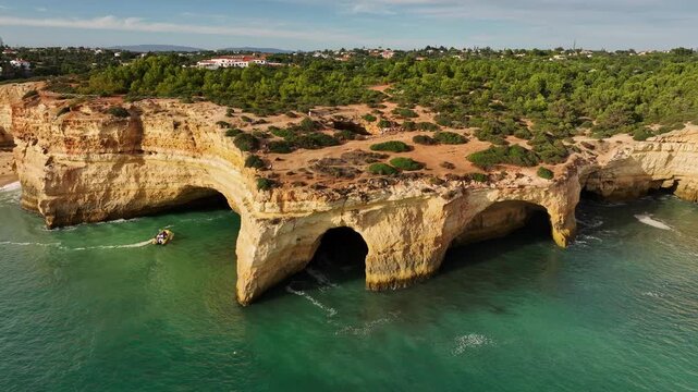 Drone view of Benagil Cave cliffs in Algarve, Portugal, with a tourist boat near limestone sea caves, turquoise Atlantic water, and rugged coast. Orbit parallax shot.