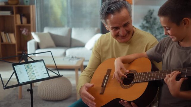 Grandfather helps his grandson learn to play guitar at home, using a tablet guitar app with chord diagrams, fingering guidance and beginner lessons in a cozy living room.