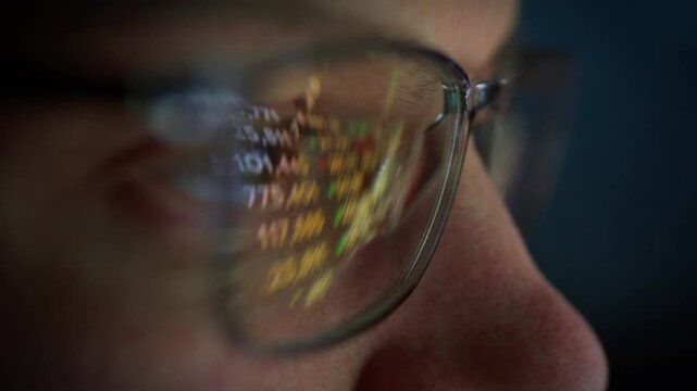 A diligent trader in glasses sits before a computer, scrutinizing live stock market screens. The glowing display reflects in his lenses, casting a faint night-time glow as he parses data