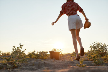 Fototapeta premium Young woman walks outdoors at sunset, wearing a striped shirt and white shorts, holding a woven hat, stepping along a sandy path. Warm golden light captures a relaxed lifestyle moment.