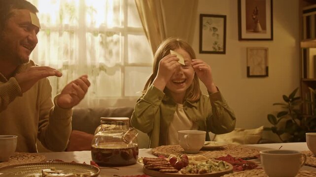 Caucasian girl playing sticky note guessing game with family at dining table while father claps. Footage fits parenting campaigns, domestic entertainment, and family bonding visuals