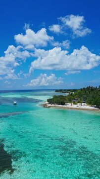 Aerial reef patterns along turquoise bay, meandering sandbars and shallow coral gardens, anchored boats near palmfringed shoreline, beachside huts and wide cloudscapes, Maldives, vertical video