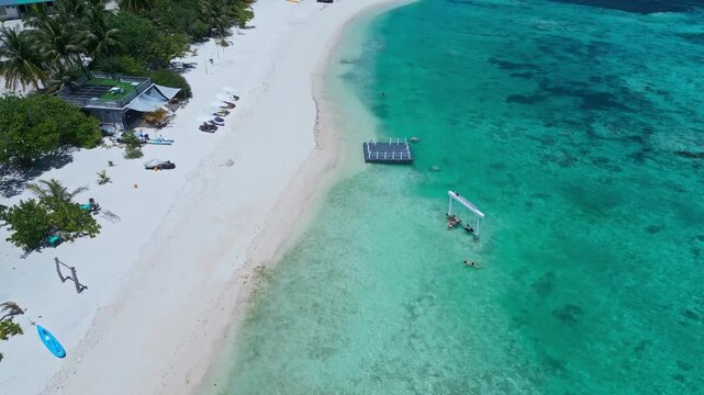 Aerial beach swing and swimmers in turquoise shallows, floating platform anchors playful activity, families wading near coral flats, white crescent sand and sunny coastal mood, Maldives,vertical video
