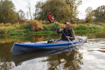 Man paddles a blue kayak with a red oar on a calm river near a grassy shore in autumn forest © WoodHunt
