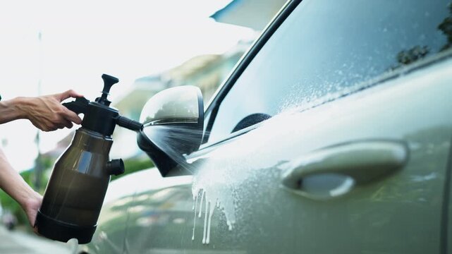 Close-up of pressure sprayer applying foam onto car door and side mirror. DIY car washing, vehicle maintenance and auto care lifestyle concept.