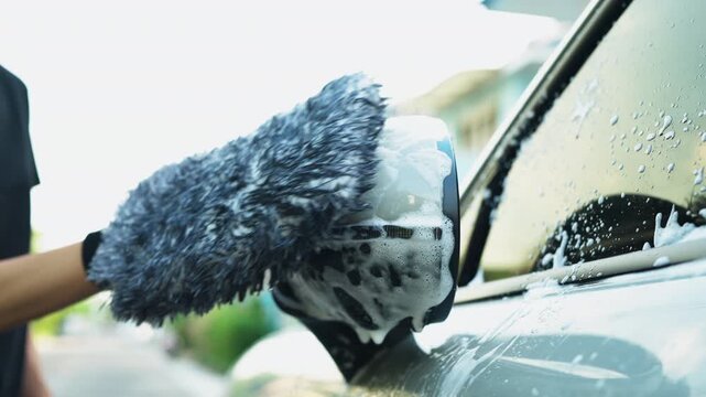 Close-up of hand using microfiber wash mitt to clean soapy car side mirror outdoors. DIY car washing, auto detailing and vehicle maintenance lifestyle concept.