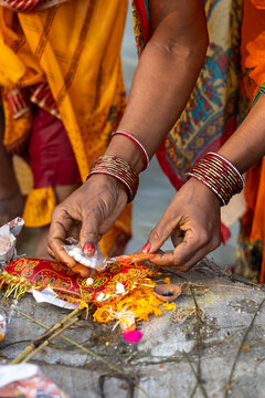 Two women with bangles arrange flowers and white sweets on a red cloth with a clay lamp at the Maha Kumbh Mela, Prayagraj, India