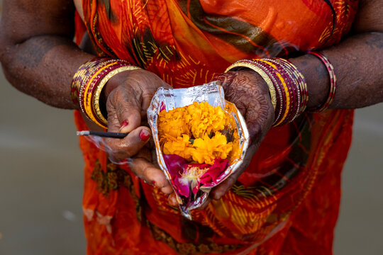 Woman in an orange sari with henna hands holds a foil packet of marigolds and an incense stick at the Maha Kumbh Mela, Prayagraj, India