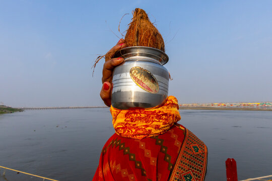 Hand holding a steel pot topped with a coconut hiding a face by the river with colorful tents at the Maha Kumbh Mela, Prayagraj, India