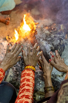 Close view of elderly women hands with red nail polish and bangles warming over a glowing fire at night, Prayagraj, India