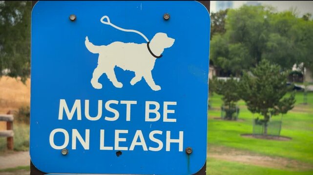Blue and white sign of a caricature of a dog on a leash, with the words "MUST BE ON LEASH" demonstrating all dogs needed to be on leash while in this public space. Trees and grass in background.