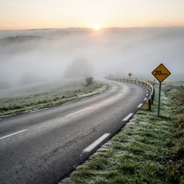 Winding asphalt road through a misty, frost-covered landscape at sunrise with a yellow 20 km/h speed limit warning sign on the roadside