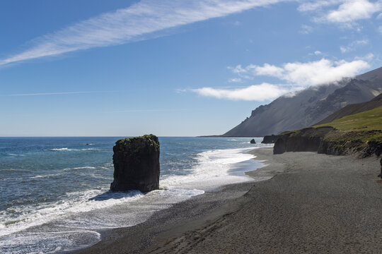 Stapinn sea stack at Laekjavik black sand beach in Iceland. White surf curves along the rugged coast beneath steep mountain slopes, grassy cliffs and blue summer sky.