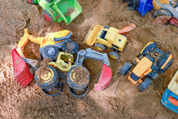 Colorful toy construction vehicles including dump trucks, bulldozers and tractors scattered in a sandy playground, outdoor play, childhood, imagination and learning through play © cgdeaw