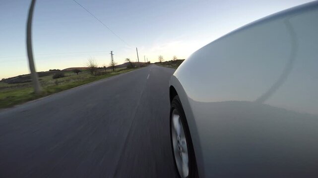Wide angle fender mount camera view of a silver car driving fast along a straight countryside road. Low action perspective showcasing asphalt highway, road markings and landscape at late afternoon.