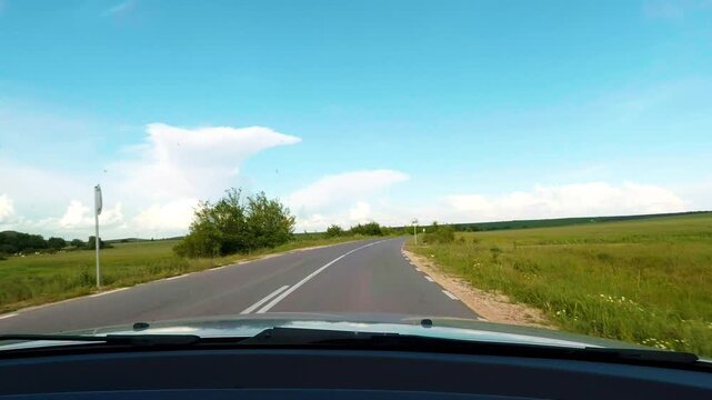 Windshield dashcam view with car hood driving along an open countryside road through green fields. Beautiful road trip on a sunny day under a blue sky with soft white clouds in Bulgaria.