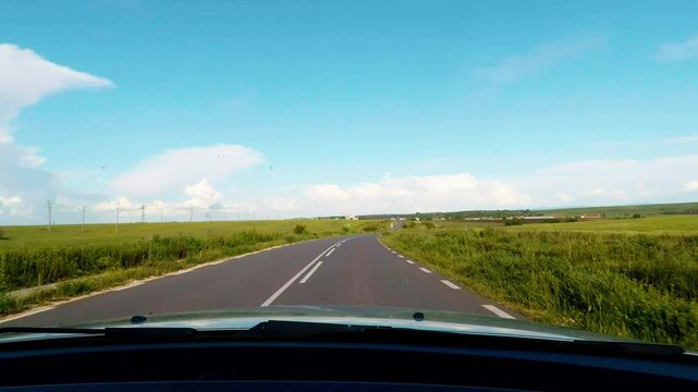 Windshield dashcam view with car hood driving along an open countryside road through green fields. Beautiful road trip on a sunny day under a blue sky with soft white clouds in Bulgaria.