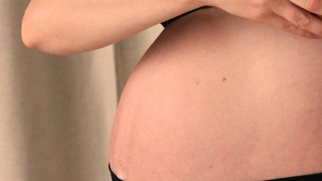 Pregnant woman gently rolls a spiky rubber massage ball across her bare belly and side, using the textured surface for myofascial release and calming daily prenatal self care wellness routine at home