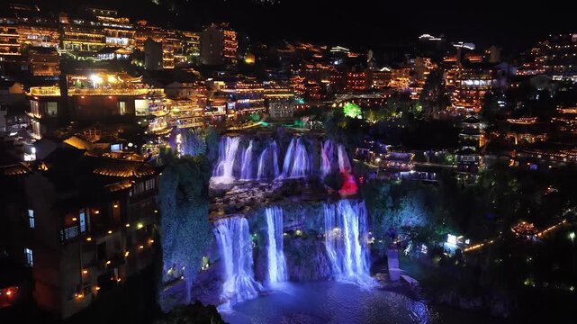 Night push-in toward the blue and purple lit Furong waterfall framed by red-lit stilt houses on the cliff and the glowing old town, Furong ancient town, Hunan, China