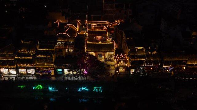 Slow push-in toward a historic hall outlined in golden lights on the riverbank at night, neon characters and people on the promenade, Fenghuang, Hunan, China