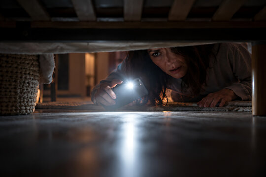Frightened woman using smartphone flashlight to look under bed at night