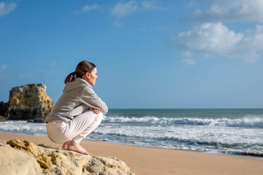 Woman in Grey Hoodie Sitting on Beach Rock Looking Out to Sea