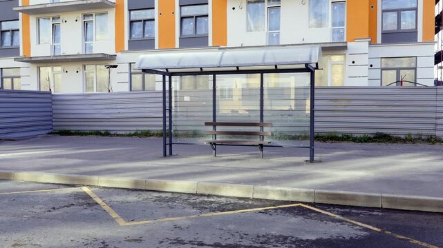 Empty contemporary bus stop with metal frame, translucent canopy, and wooden bench. Set on a paved platform with a low curb and yellow road markings in the foreground