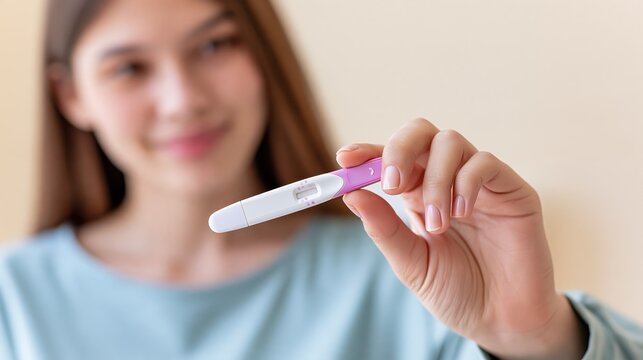 A young woman holding a pregnancy test in her hand while smiling at the camera in soft lighting