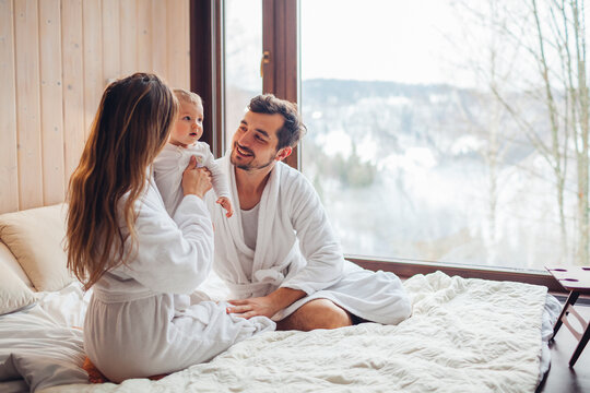 Happy family playing on bed enjoying snowy mountains landscape from hotel window. Mom and dad having fun with toddler baby girl