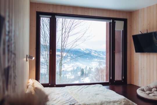 Interior of cabin house with winter landscape in mountains. Bedroom with panoramic window and snowy forest view