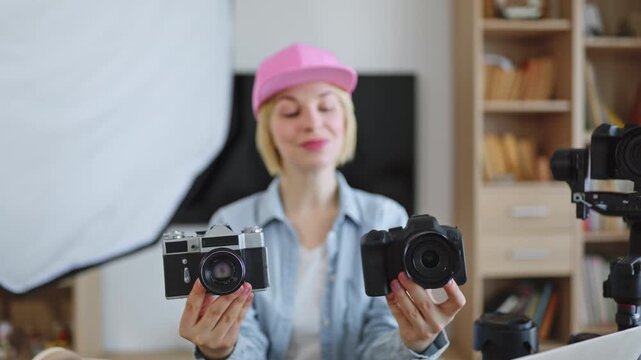 A young woman wearing a pink cap holds two different cameras, a vintage film camera and a modern digital camera, in front of a bookshelf