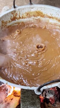 thick brown dodol candy cooking in metal pot over open fire, stirring sticky traditional asian dessert mixture with wooden paddle in close-up, slow motion in 60fps