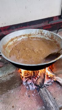 thick brown dodol mixture in large metal wok over open wood fire, bubbling sweet sticky candy dessert with rising steam, outdoor cooking scene, traditional asian culinary preparation